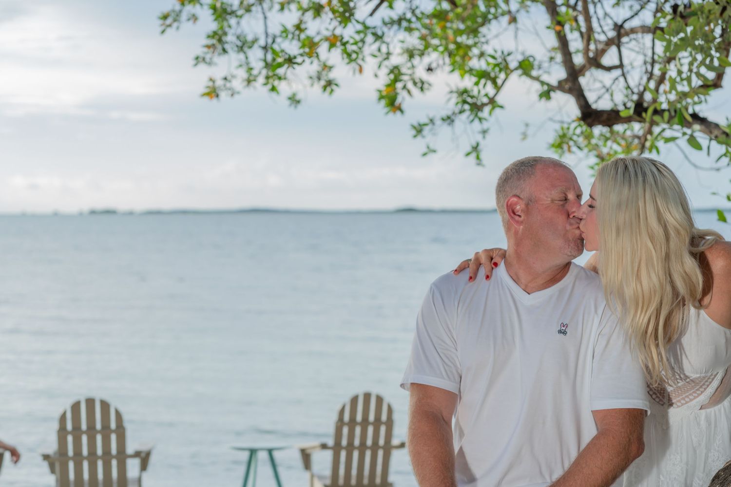 A couple shares a romantic moment on the waterfront with white Adirondack chairs and ocean views in the background.