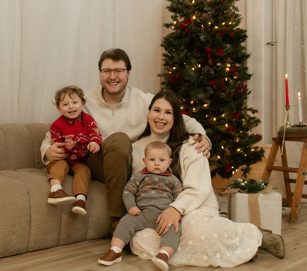 A family sits together on a beige couch in front of a decorated Christmas tree with candles nearby.