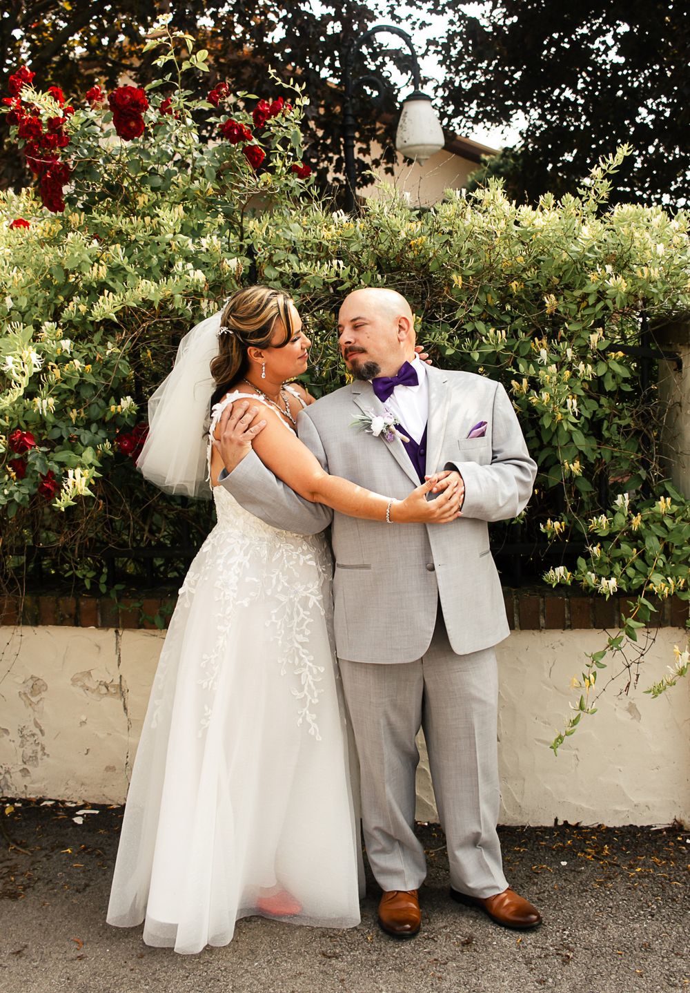 A couple in wedding attire shares a romantic moment looking at a phone in a garden setting.
