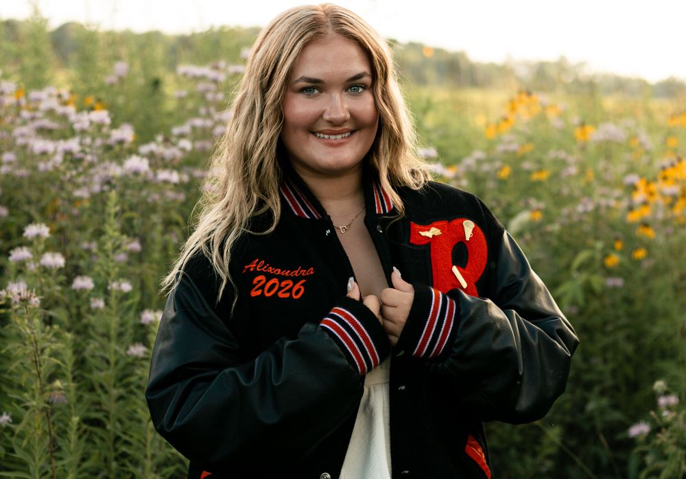 A person in a black and red varsity jacket poses in a sunny wildflower field during golden hour.