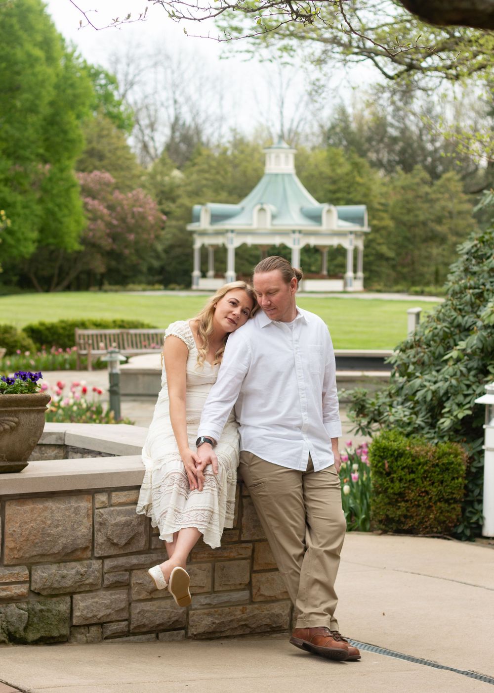 Couple poses romantically in a lush garden with a gazebo backdrop, woman in white dress leaning on stone wall.