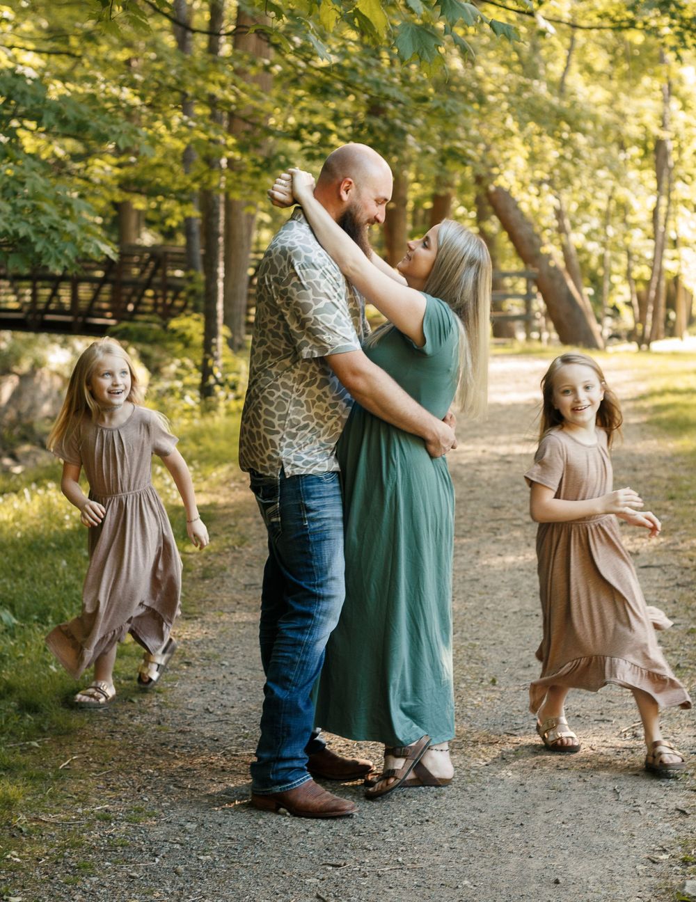 A couple embraces on a wooded path while two young girls in flowy dresses play around them during a family photo session.
