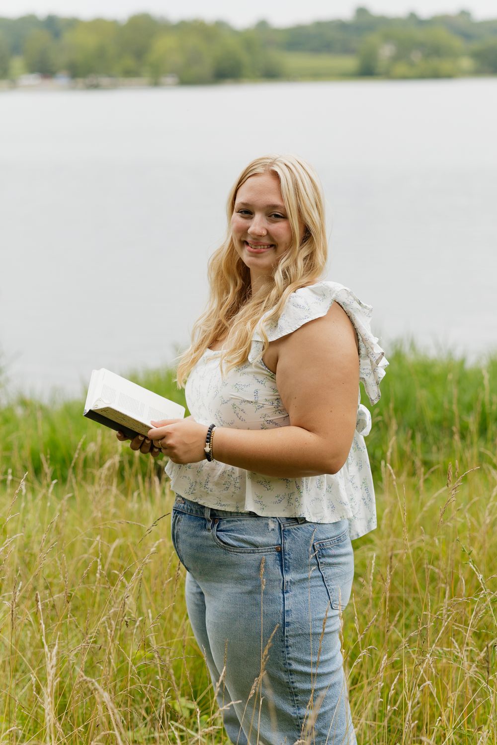 Person in casual attire reading outdoors by a lake surrounded by tall grass on a sunny day.