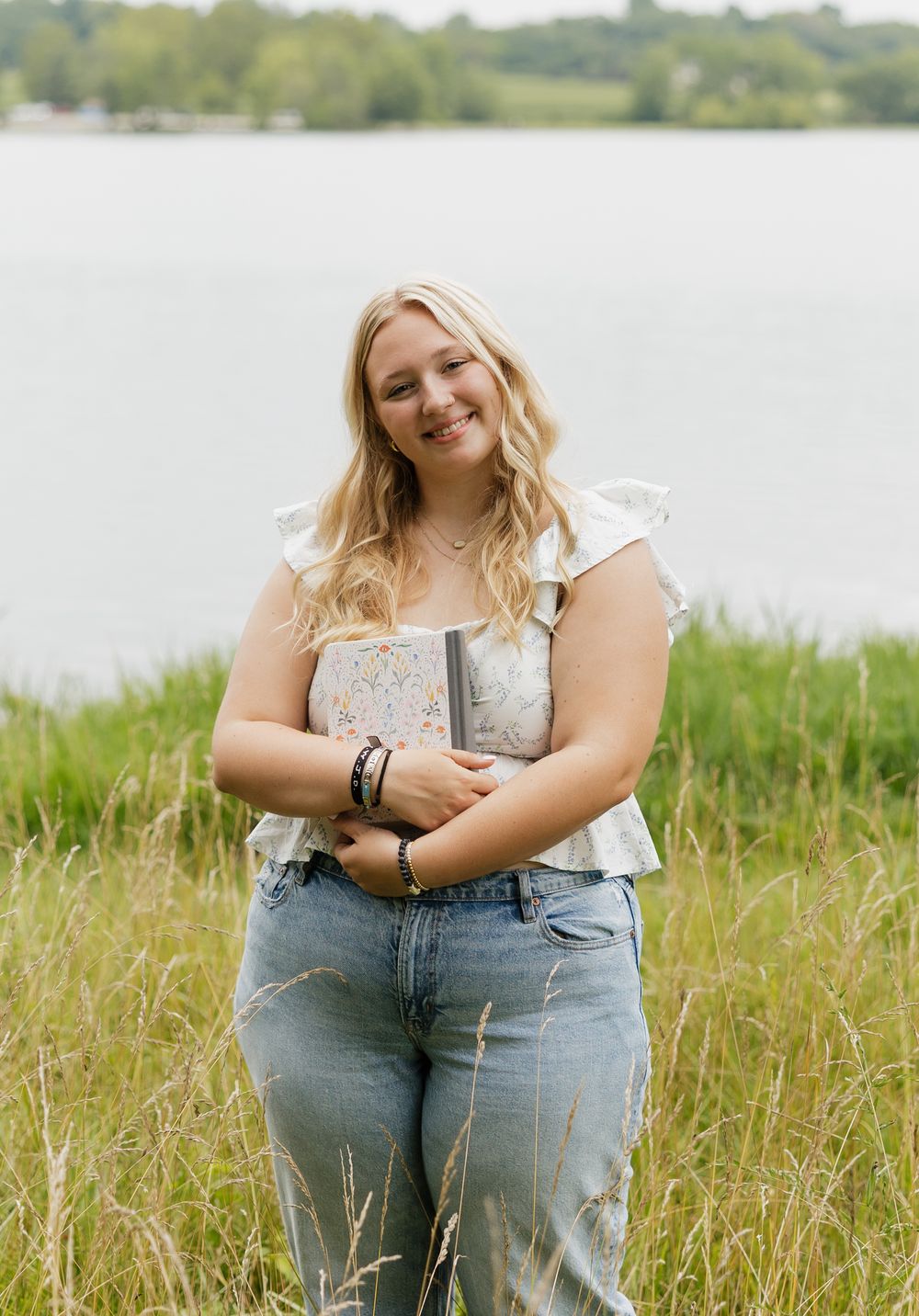 A figure sits in tall grass by a lake wearing jeans and a light colored top on a sunny summer day.