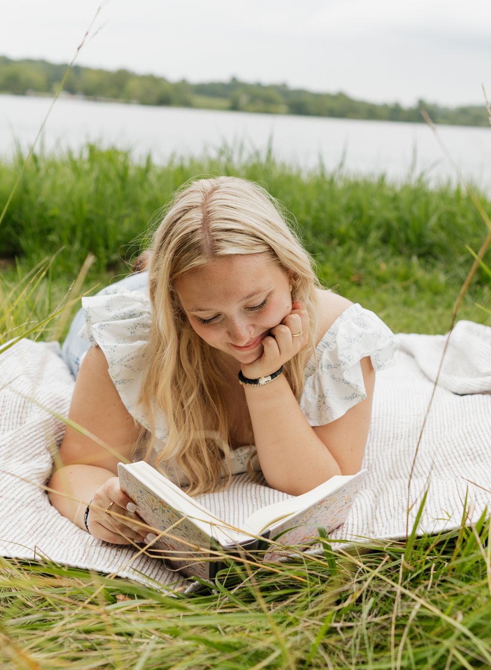 Person lying on grass reading a book on a white blanket near a lake on a sunny day.