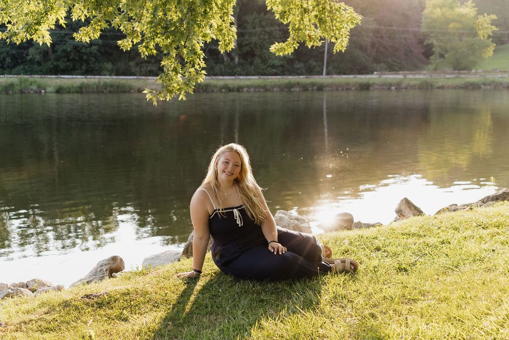 A person relaxes by a serene lake on a grassy bank with golden sunlight filtering through trees.