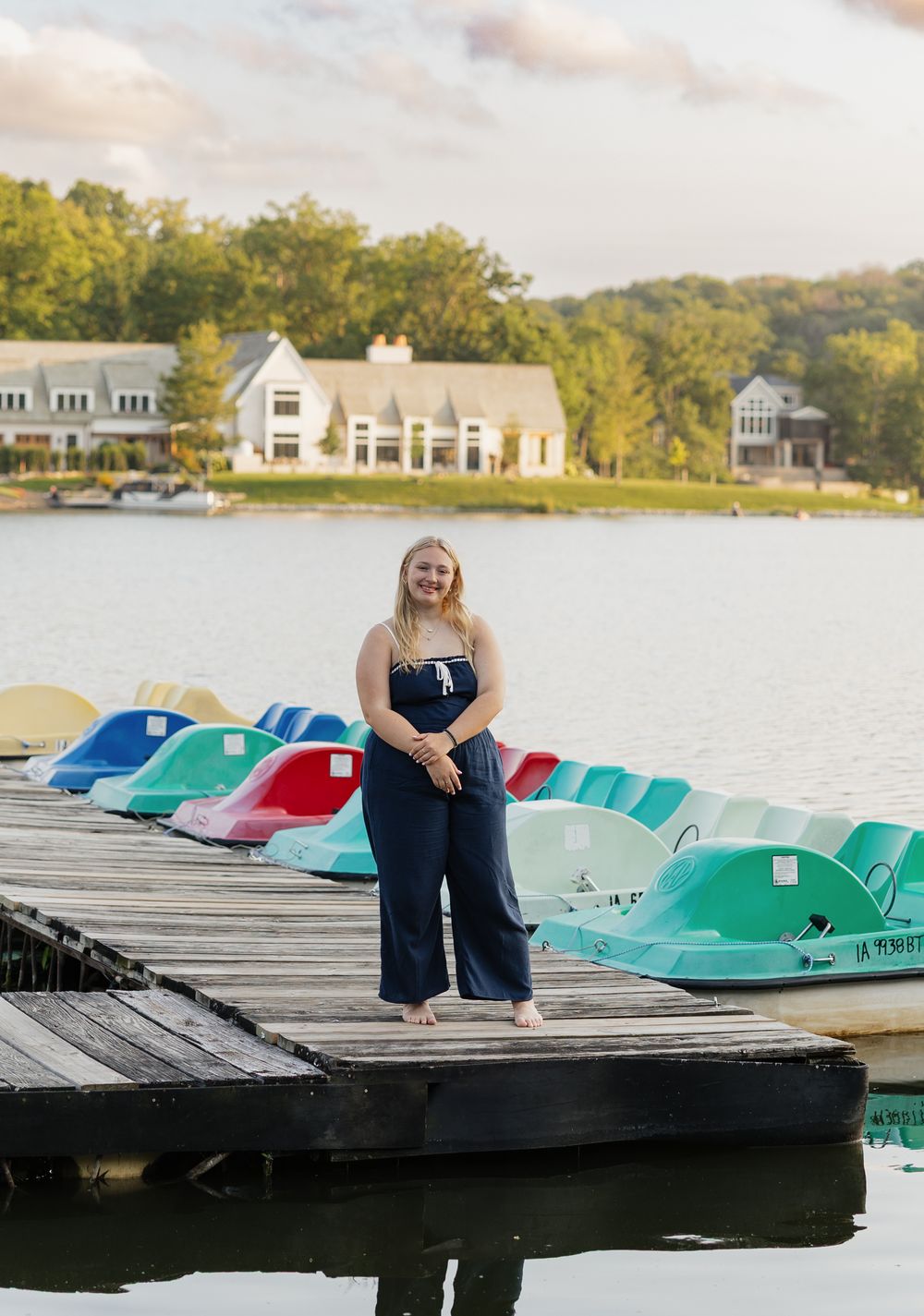 A row of colorful paddle boats line the wooden dock on a serene lake with houses visible in the background.