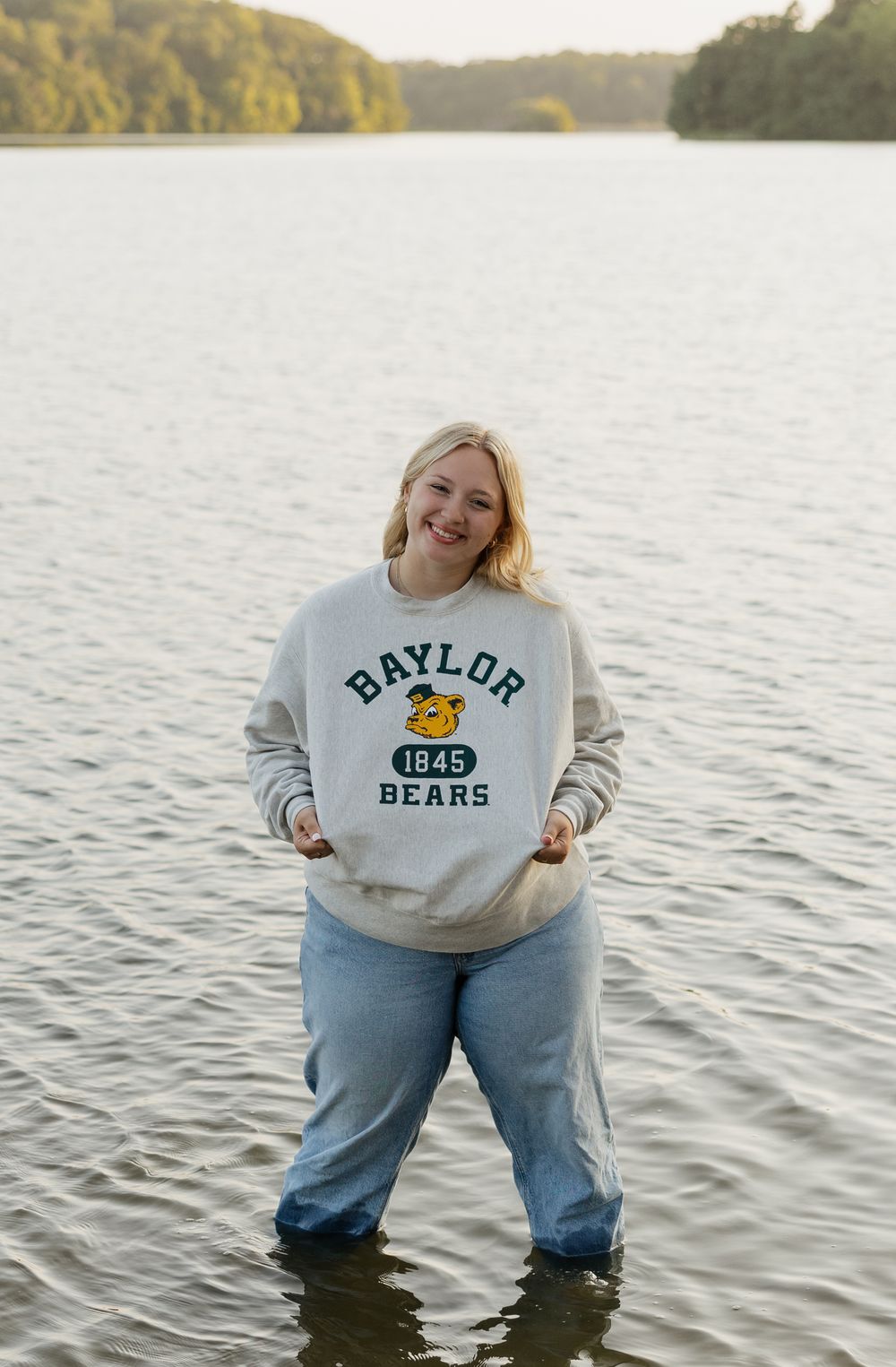 Person wearing a Baylor Bears sweatshirt standing in lake water during golden hour.