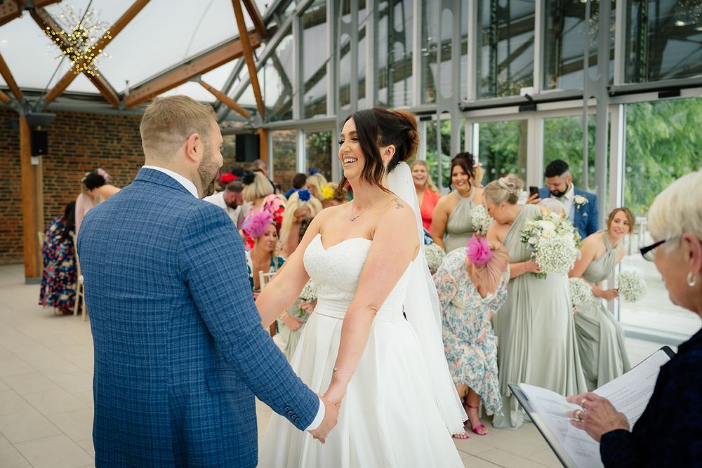 Wedding ceremony guests seated in rows of white chairs inside a bright, modern glass-walled venue with natural lighting.