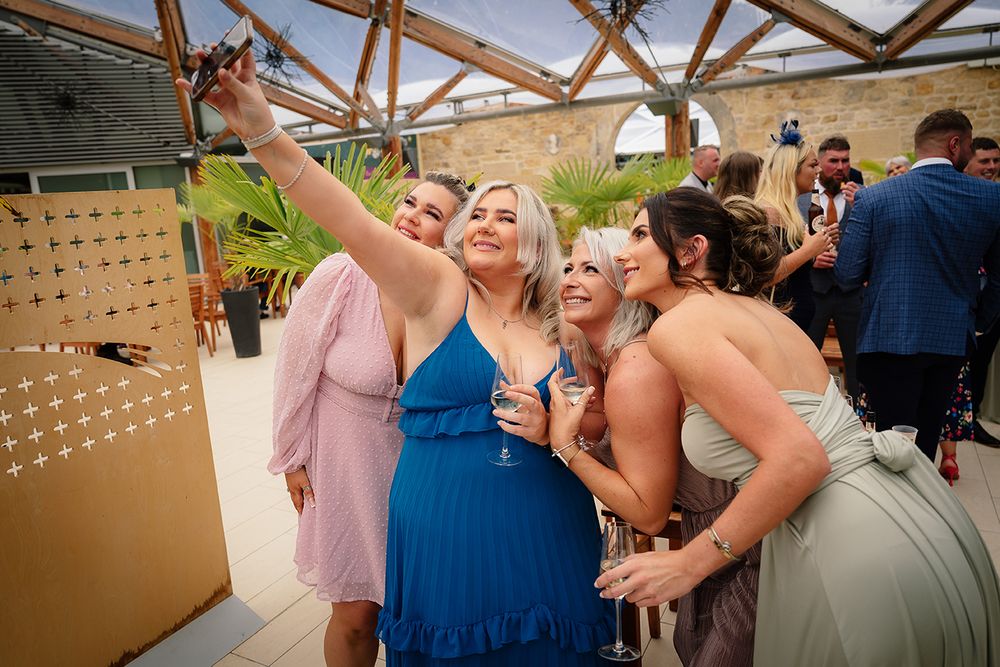 A group of friends in formal attire taking a selfie at an outdoor event under a wooden pergola structure.
