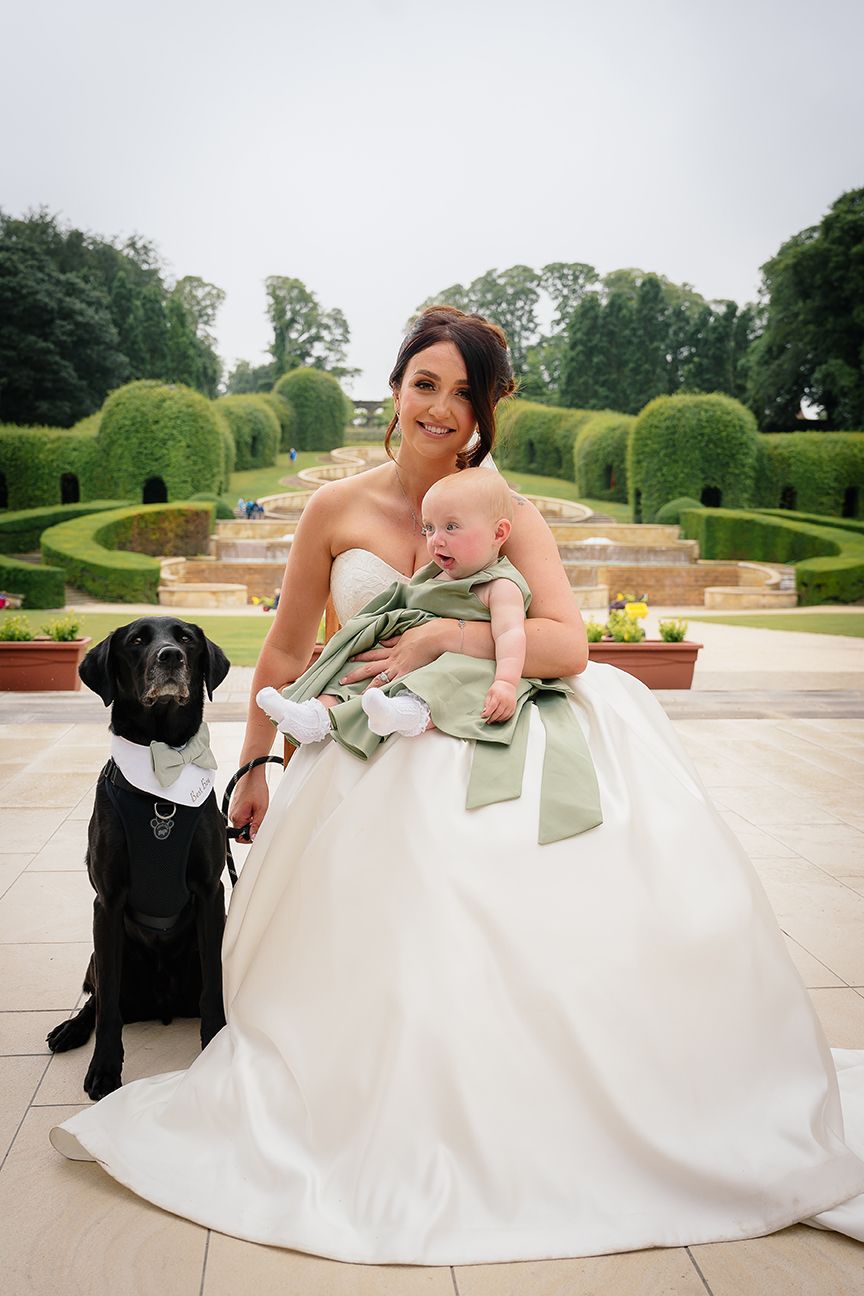A bride in a white gown sits on stone steps with a baby in green and a black Labrador wearing a bow tie at a garden venue.