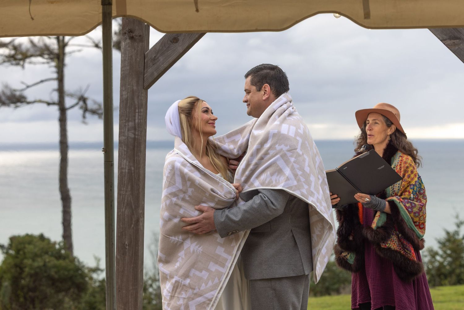 Couple wrapped in blankets embrace during outdoor ceremony while officiant stands nearby on cloudy day.