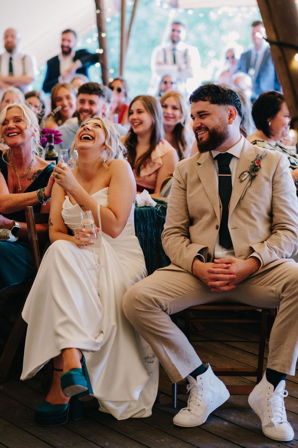 A couple in wedding attire share a joyful moment of laughter while seated among guests during a reception celebration.