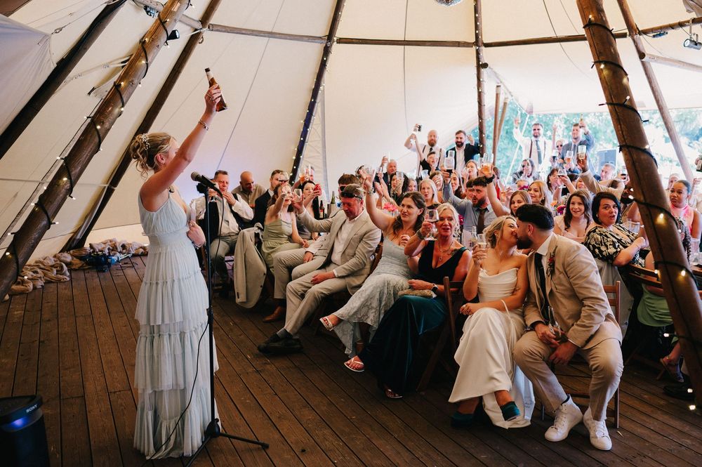 Wedding guests cheer and celebrate inside a large tipi-style tent during a festive reception celebration.