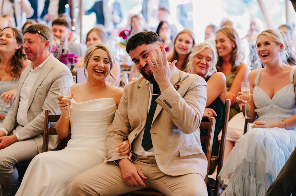 Wedding attendees laugh and react emotionally during speeches at an outdoor tent reception.