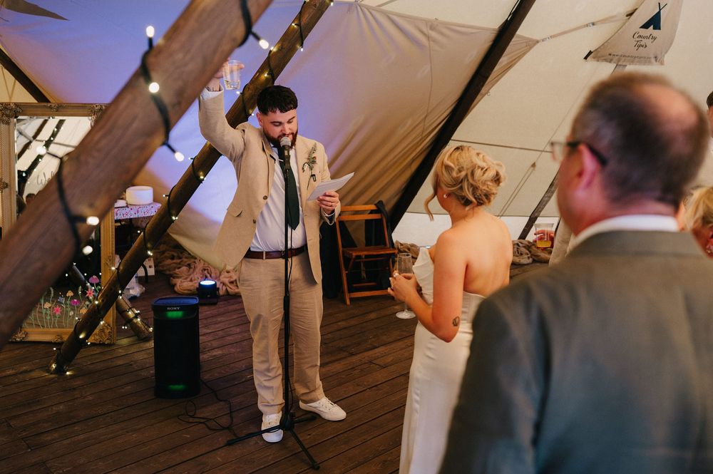 A speaker entertains guests during a wedding reception in a tent venue with string lights.