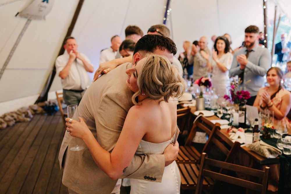 A tender embrace shared during a wedding reception as guests applaud in the background under a tent setting.