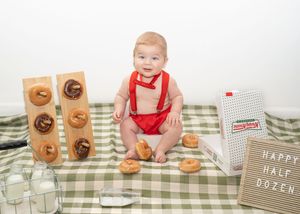 Baby in red suspenders sits on checkered blanket with donuts and 'Happy Half Dozen' sign for 6-month milestone photo.