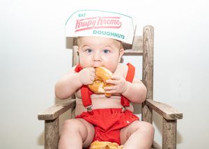 A small child in a red outfit sits in a wooden chair eating a doughnut.