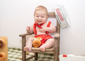 Baby in red overalls sits in rustic wooden chair during photo session with teddy bear and holiday decor.