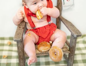 Baby in red suspender outfit sits in wooden chair with donuts scattered around on green checkered blanket.