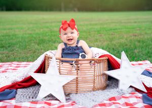A baby in a red bow and navy outfit sits in a wooden crate with patriotic decor on a picnic blanket.