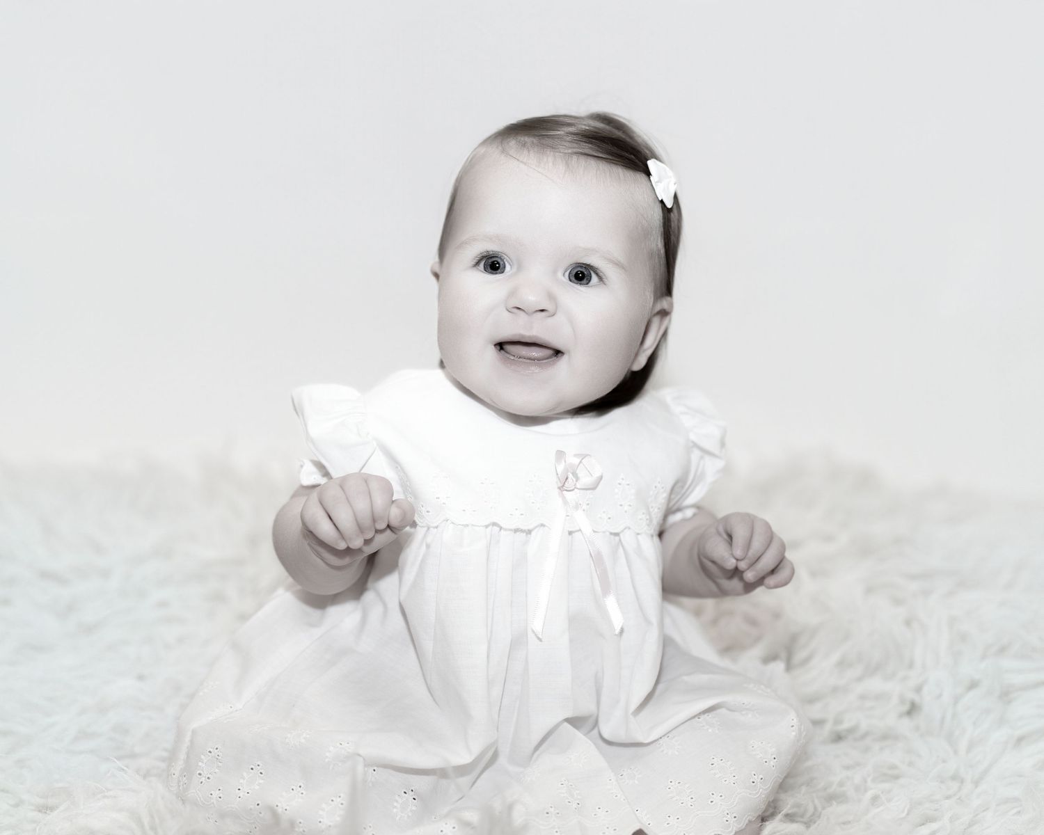 Baby wearing white dress smiles while sitting on soft furry blanket in black and white portrait.