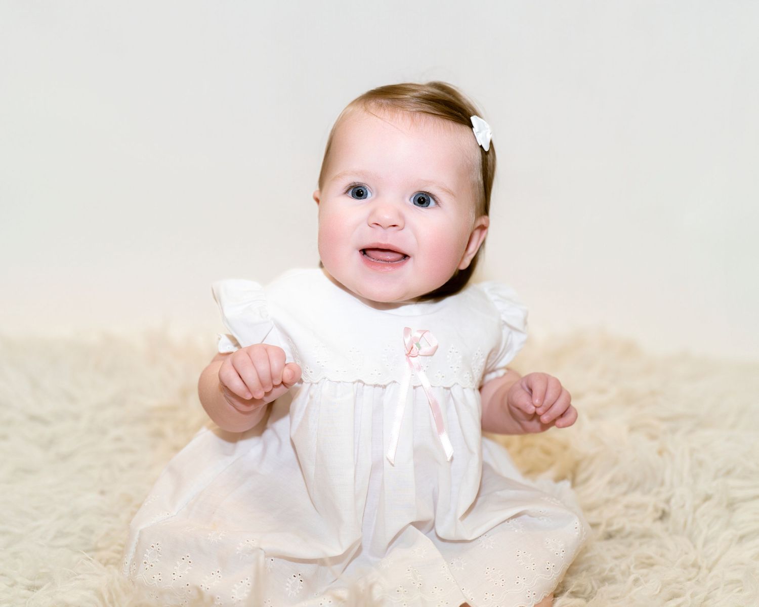 A baby in a white dress sits and smiles on a light colored carpet.
