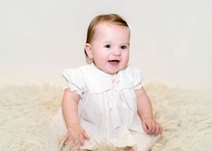 A baby in a white dress smiles while sitting on a cream-colored carpet.