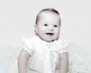 Sweet baby wearing white dress smiles while sitting on soft white backdrop in classic portrait style.