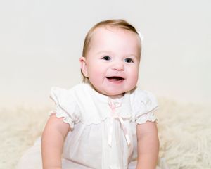 A baby wearing a white outfit smiles while sitting on a cream-colored blanket in a series of connected photos.