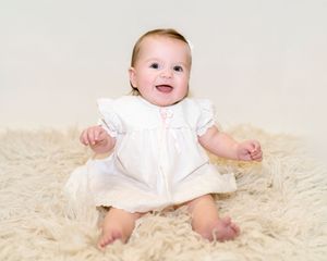 Adorable baby in white dress sits on cream colored carpet during professional photo session.