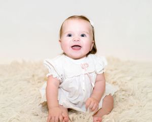 A baby in a white dress smiles while laying on a soft cream-colored carpet.