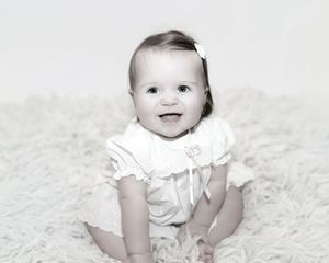 Baby in white dress sits on plush white carpet during studio photo session.