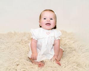Happy baby in white dress sitting on beige fluffy carpet in studio setting.