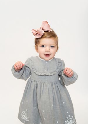 A baby wearing a gray ruffle dress with embroidered floral details along the hemline poses in three different positions.