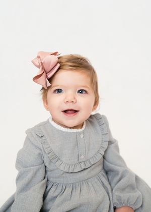 A baby wearing a gray ruffled dress with pink hair bows poses against a white background.