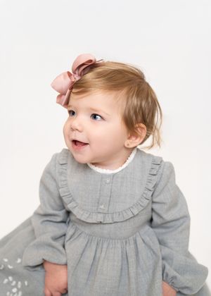 Baby in gray dress with ruffled collar smiles while looking up against white backdrop.