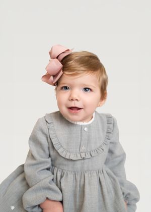 Baby wearing a gray ruffled dress and pink bow against white background smiles sweetly.