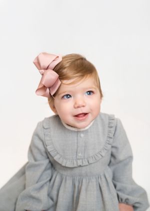 Sweet baby in vintage-style gray dress with pink bow accessory against white backdrop.