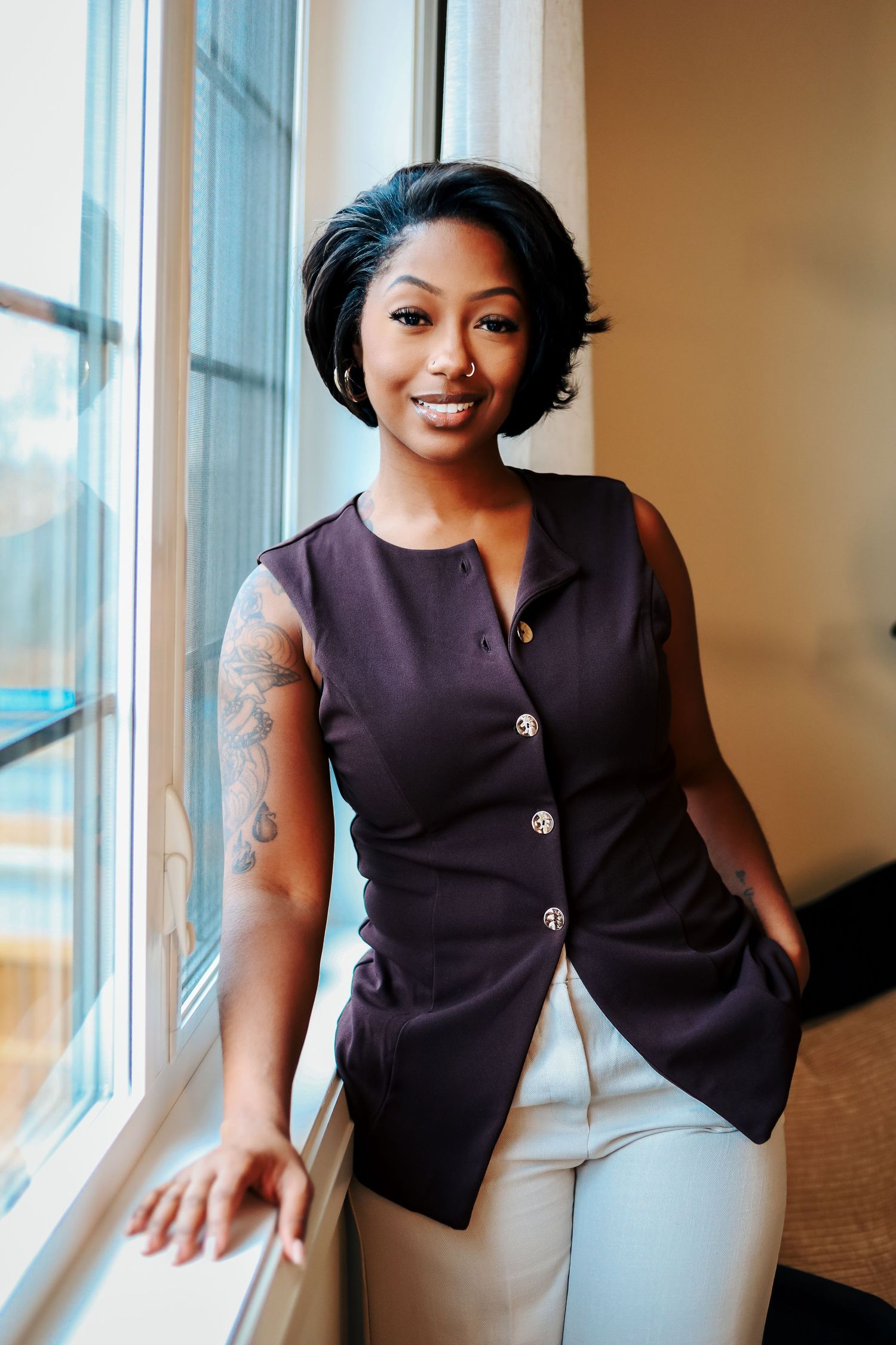A woman in purple vest and white pants poses by window, showcasing tattoos and short curly hair. Headshots and branding photos near edmonton