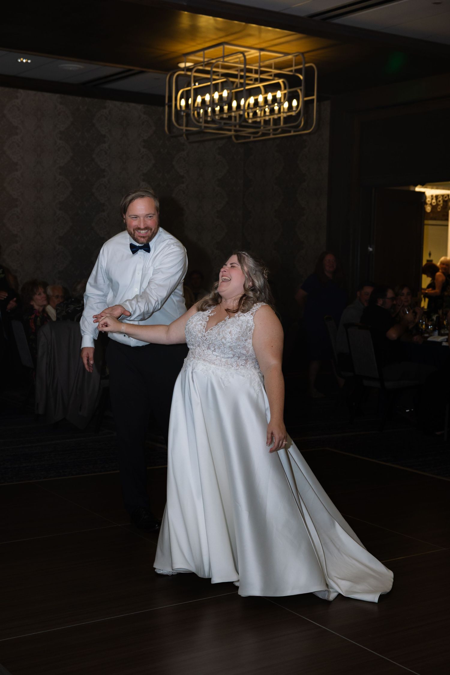 A couple shares a romantic dance moment on a dimly lit reception floor under elegant chandelier lighting.