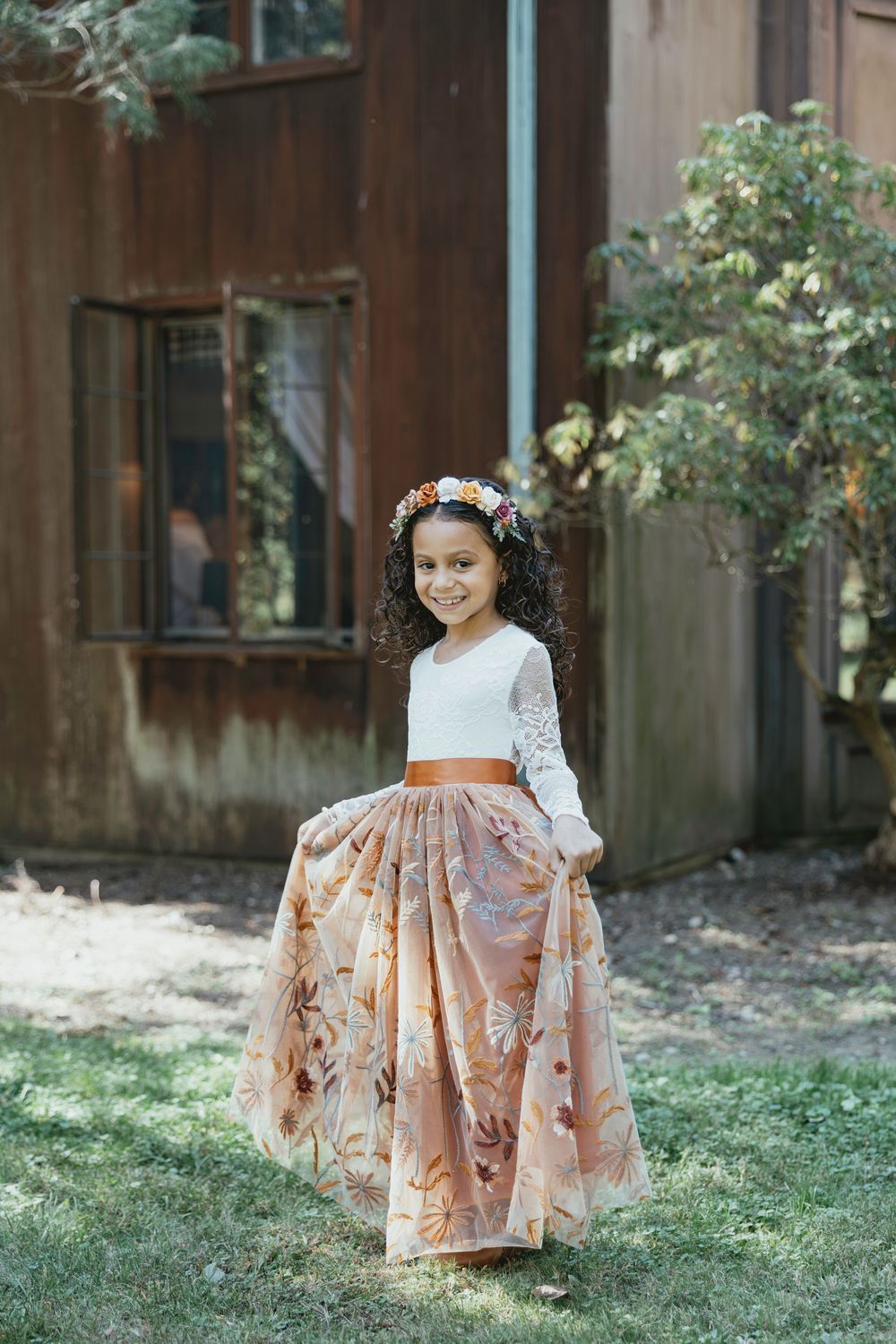 A young girl wearing a white lace dress with an orange sash and floral skirt stands in a garden setting.