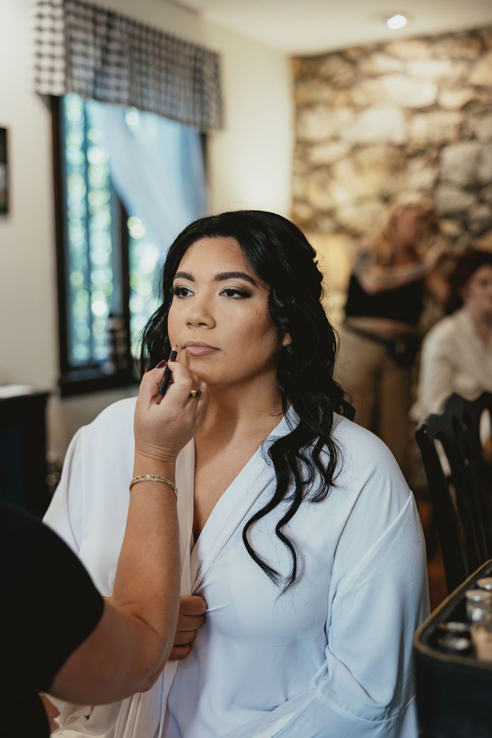 A person in a white robe sits while getting makeup done during wedding preparations.