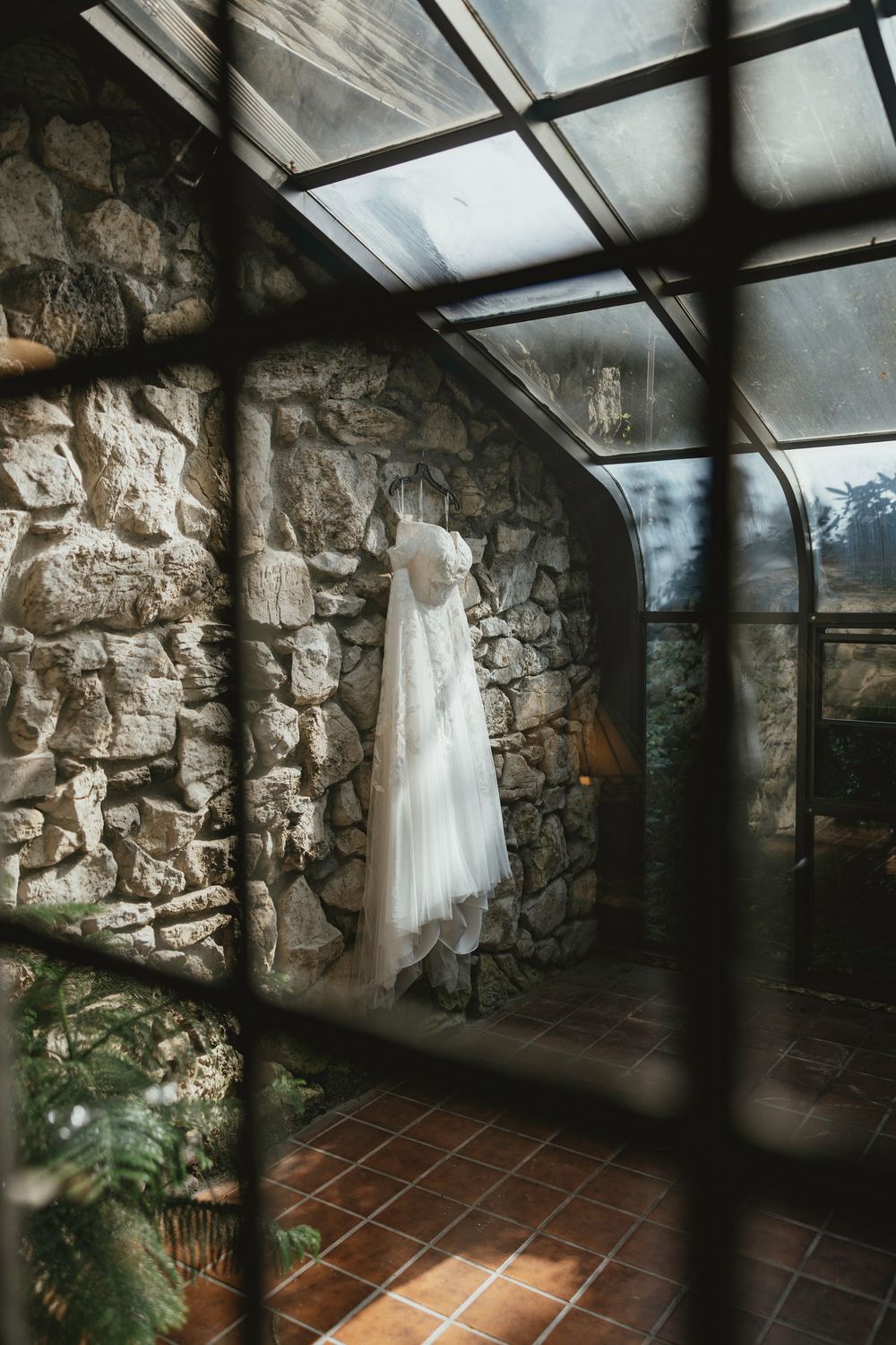 A white wedding dress hangs against a rustic stone wall in a sunlit room with terracotta floors and glass ceiling.