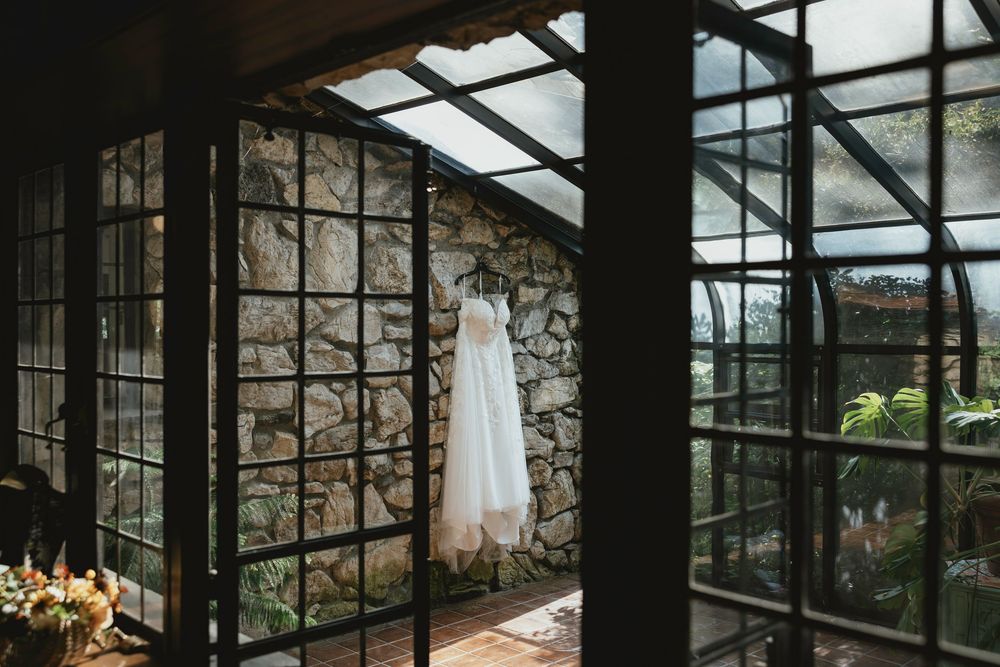A rustic greenhouse with stone walls and glass panels showcases a wedding dress hanging in natural light.