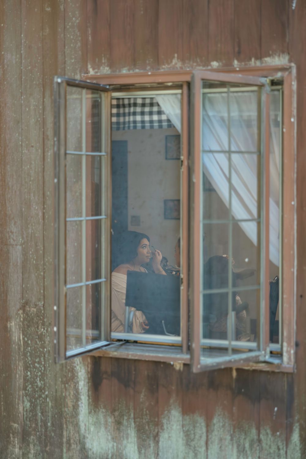 Through an open rustic window frame, a figure sits quietly in a dimly lit interior with gingham curtains visible.