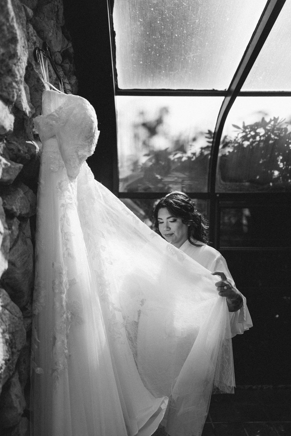 A delicate white wedding dress hangs by a window as someone admires its flowing fabric in black and white photography.