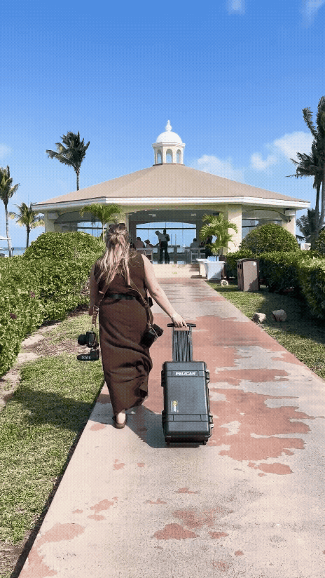 A person pulling luggage along a palm-lined walkway leading to a white gazebo in a tropical resort setting.