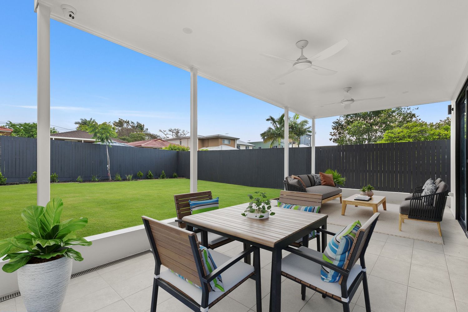 Covered outdoor alfresco area with dining table, lounge seating, and lush green lawn.
